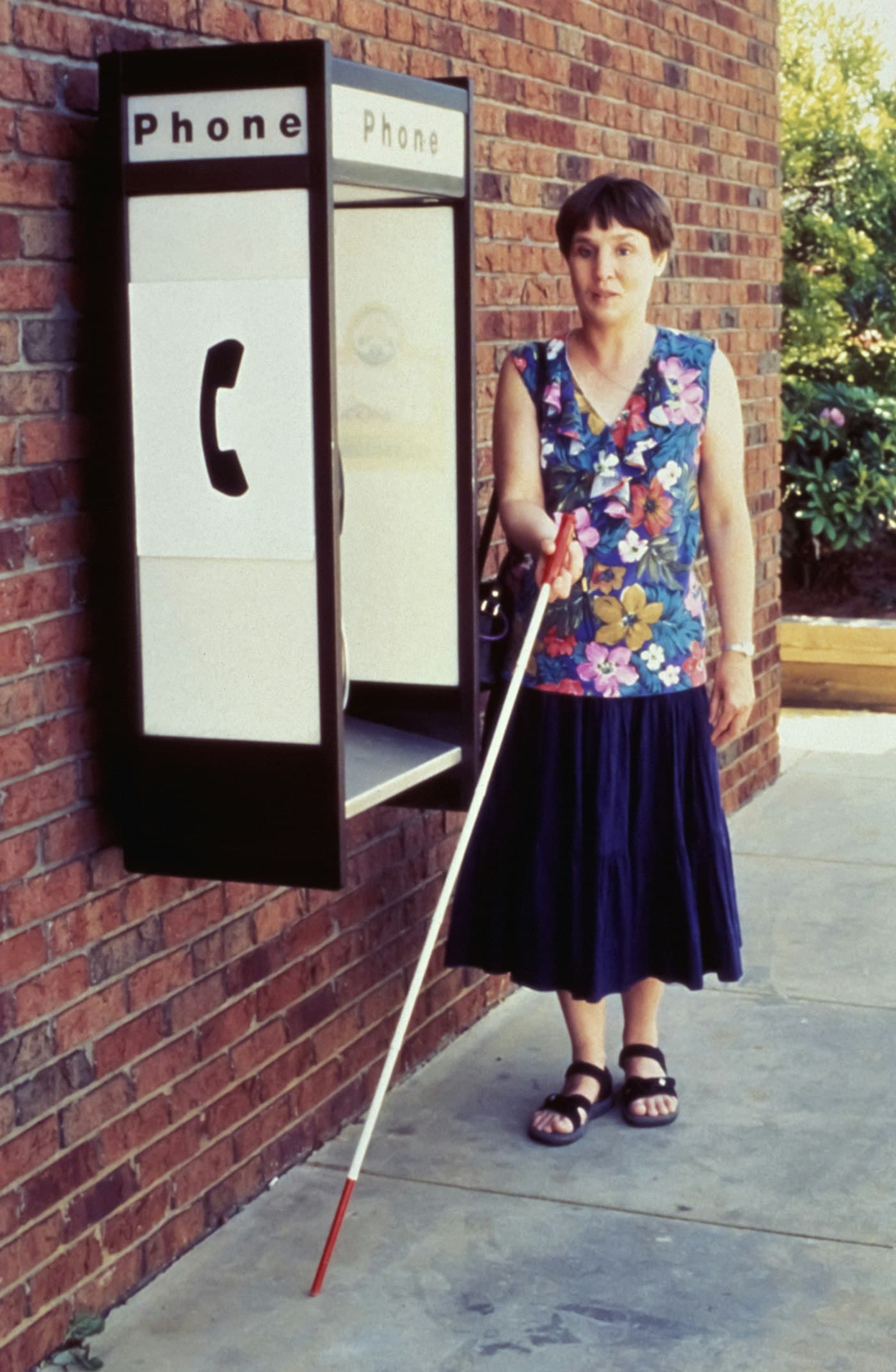 A visually impaired woman with a white cane at a public phone booth, representing accessibility and disability rights in public life