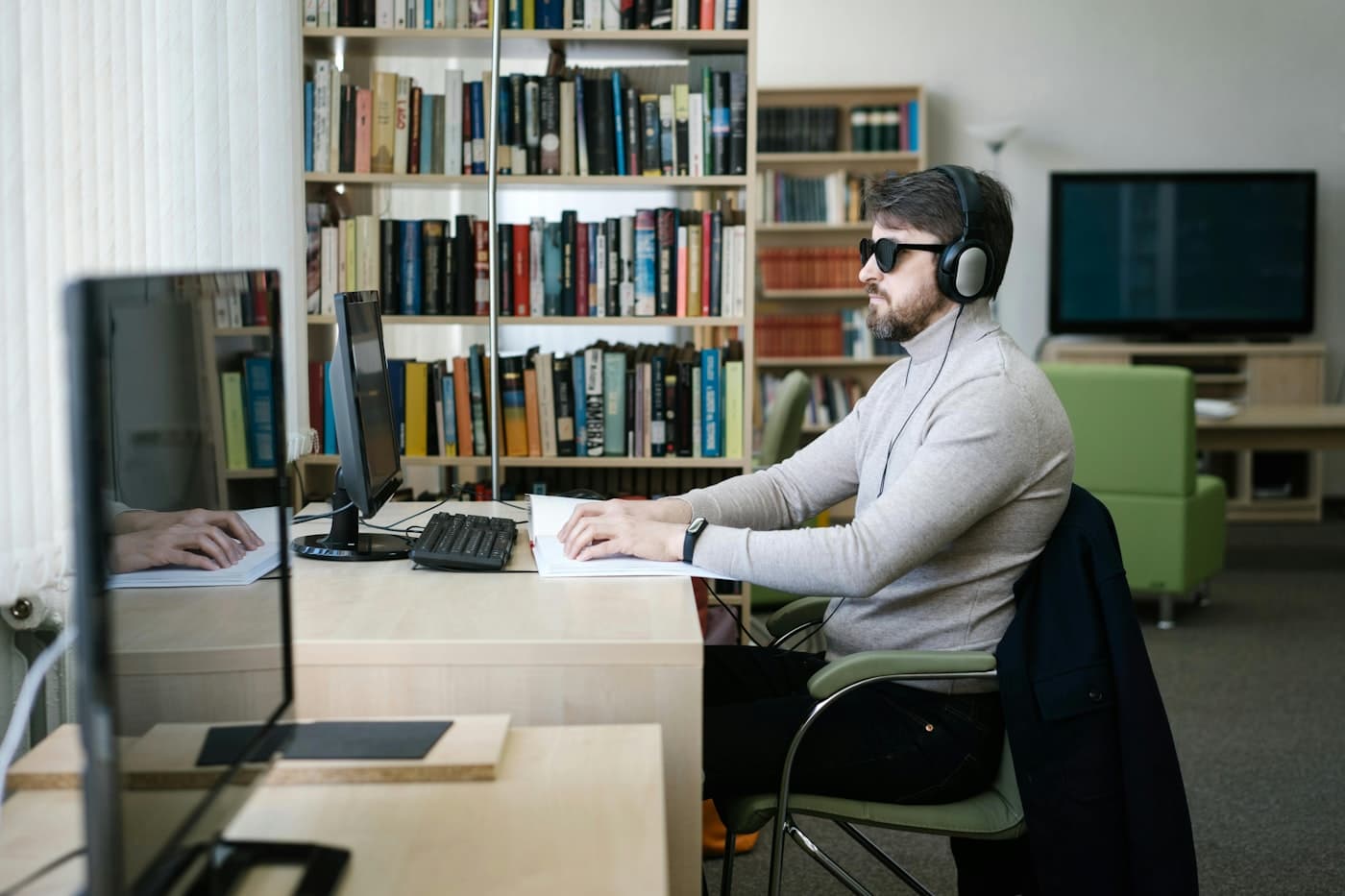 A visually impaired man using headphones, a computer, and braille materials for learning