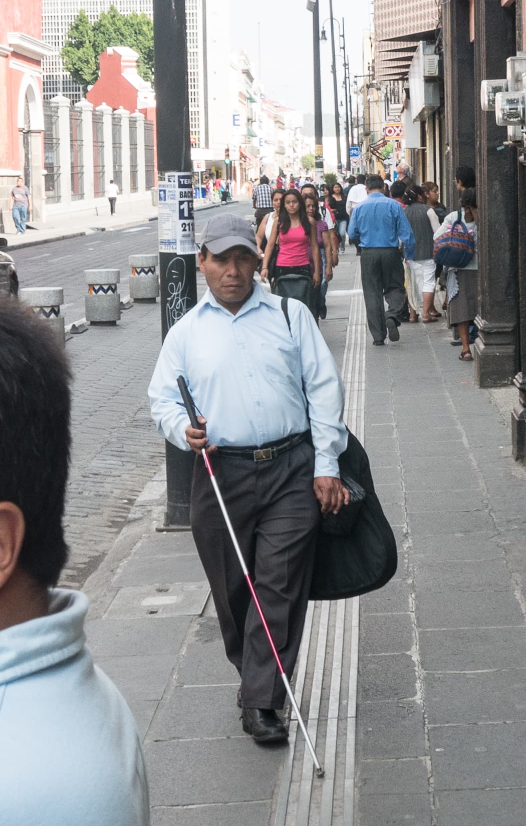 A blind man walking with a white cane on tactile paving while carrying a guitar case, representing enterprise and livelihoods